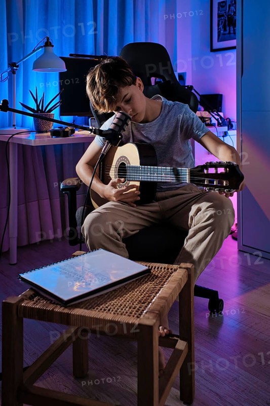 A young boy plays guitar in a cozy, dimly lit room with a microphone. The ambiance includes blue and purple lighting, creating a creative and inspiring atmosphere at home