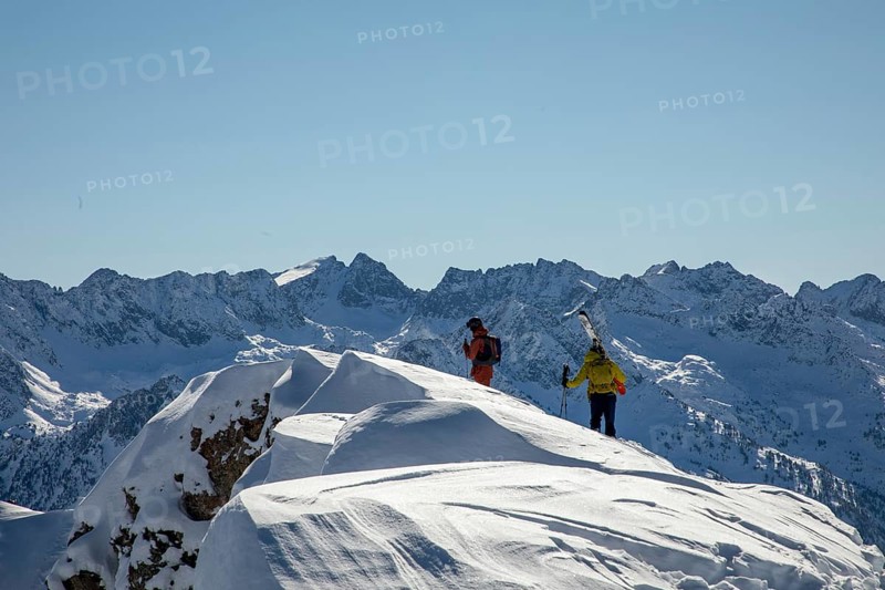 Two freeride skiers traverse a snowy ridge under a clear blue sky, surrounded by stunning mountain peaks The untouched snow hints at thrilling adventures ahead