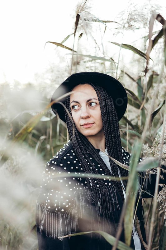 A woman stands amidst tall grasses at sunset, her braided hair gently catching the last light of the day. Her black hat adds a touch of elegance to the serene natural setting