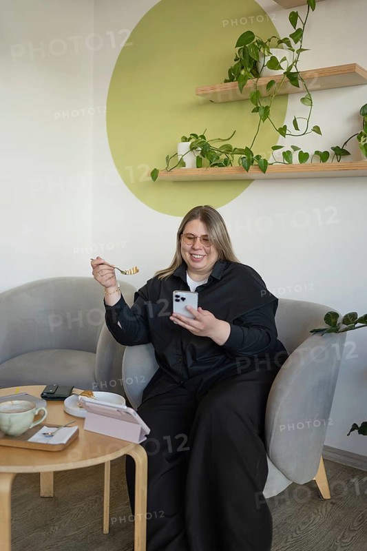 A woman in a cafe, smiling as she enjoys her snack with coffee and checks her smartphone. The cozy corner features green plants and a relaxed atmosphere