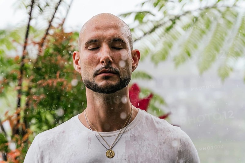 A man stands with closed eyes, soaking in the rain, surrounded by lush greenery. The scene captures a moment of mindfulness and connection with nature