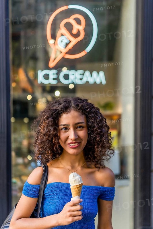 Vertical photo of a cheerful latin woman eating ice cream outside the store with neon light with ice cream shape and the logo