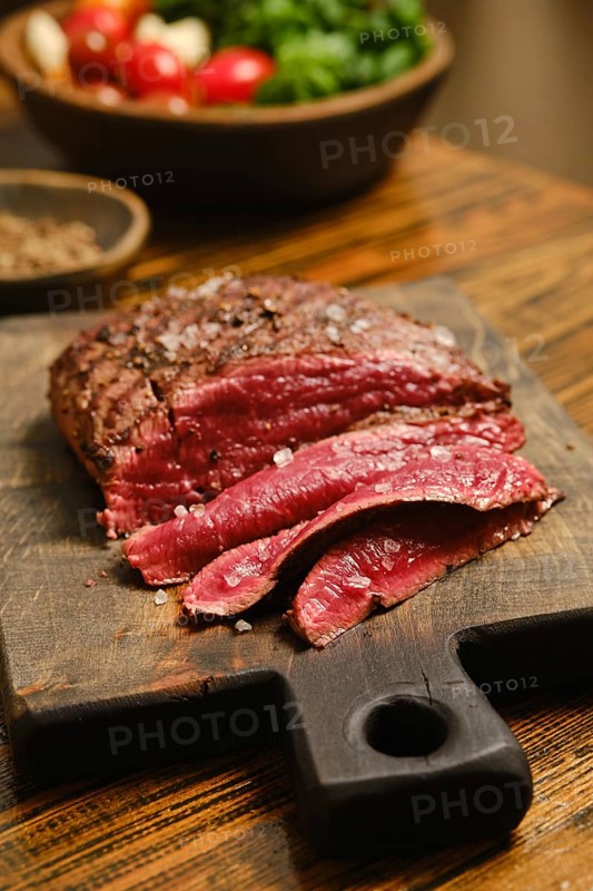 Beef steak is sliced and placed on a wooden cutting board. The meat shows a deep color and is seasoned with salt. Fresh vegetables are present in the background
