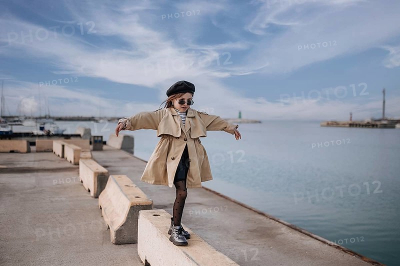 A fashionable girl balances on a concrete barrier at Civitanova's waterfront in Italy. She wears a trendy coat, beret, and sunglasses, exuding confidence under the open sky