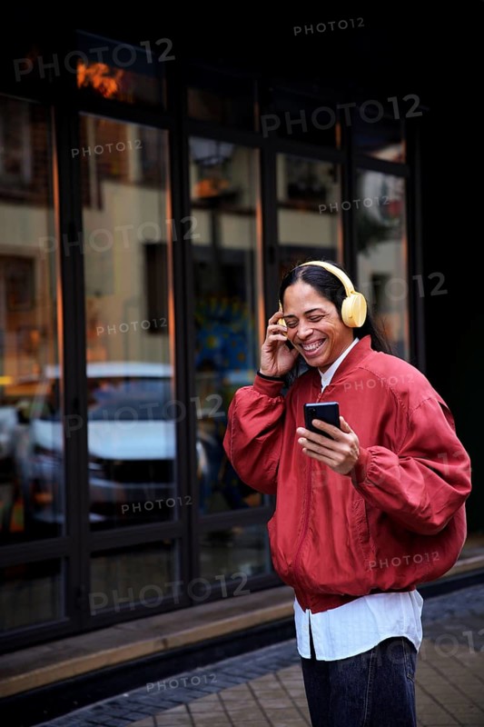A man wearing yellow headphones, holding a smartphone, and smiling while enjoying music outside. They wear a red jacket, standing by a glass building facade
