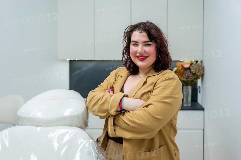 Confident female doctor smiling, standing in an aesthetic clinic room with a treatment chair, providing a welcoming and professional atmosphere for beauty and skincare services