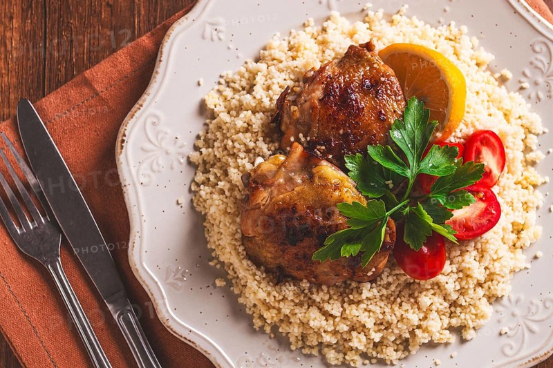 Couscous is served on a plate with pieces of chicken on top. Lemon slices and parsley decorate the dish