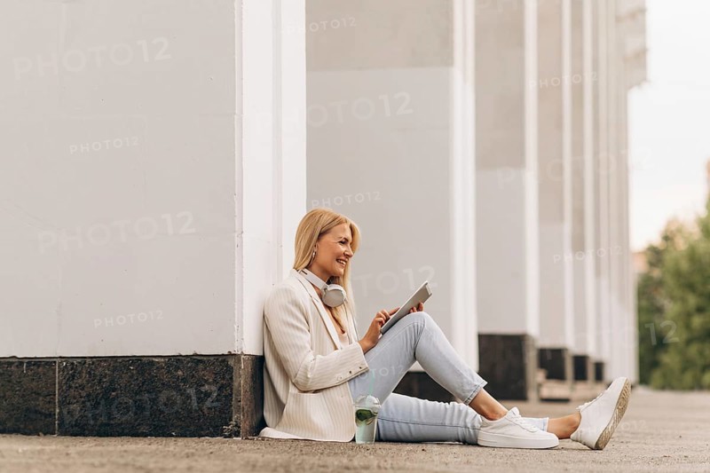 A businesswoman sits against a building, smiling while using a tablet. She wears headphones around her neck and sits casually in a relaxed, outdoor setting