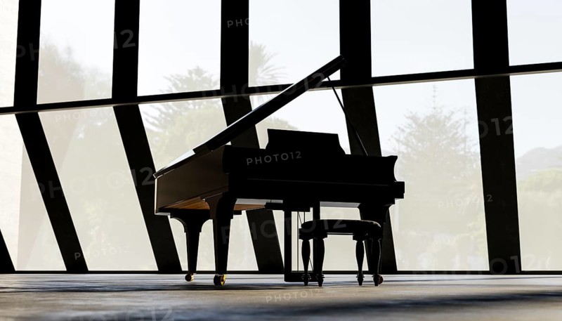 A grand piano stands in an empty, sunlit room with expansive, angular windows. The sleek silhouette and minimalist design create a sophisticated atmosphere