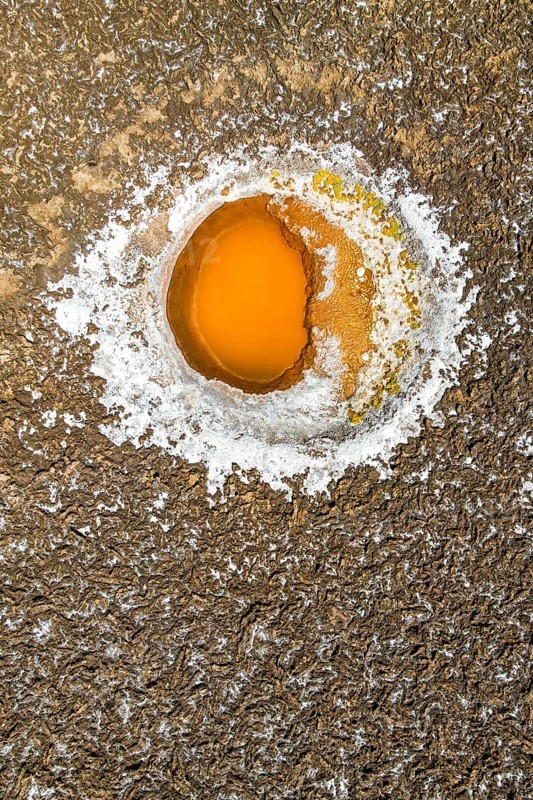 Top view aerial shot captures a striking salt pond amidst the textured landscapes of Salar de Antofalla in Catamarca, Argentina. Its vibrant orange center contrasts beautifully with the encrusted salt edges
