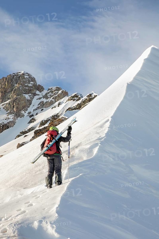 A ski tourer ascends a snow-covered ridge, skis on shoulder, against a backdrop of rugged peaks. Crisp morning light enhances the sense of adventure and exploration