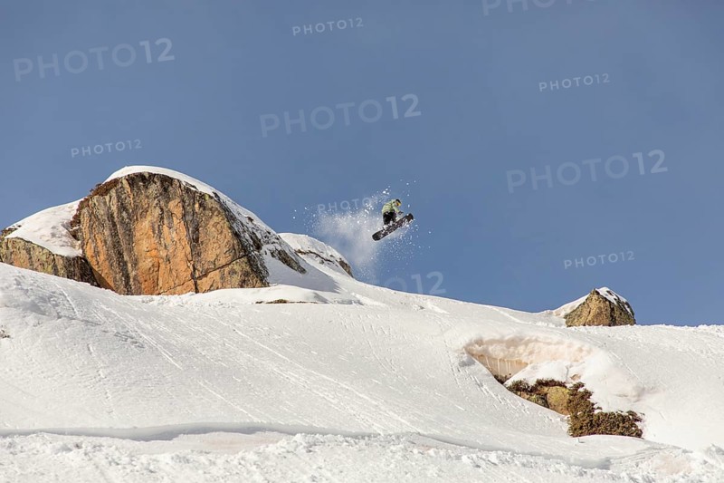 A snowboarder performs a thrilling jump off a snowy cliff, sending powder flying in the backcountry. This captures the essence of freestyle snowboarding in winter's untouched mountains