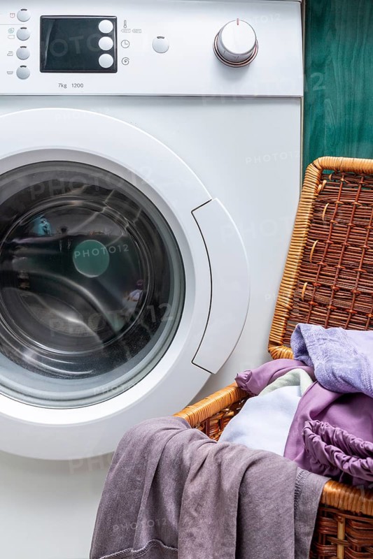 A sleek front-load washing machine stands beside a wicker laundry basket filled with soft pastel clothes, presenting a clean and modern laundry room setting
