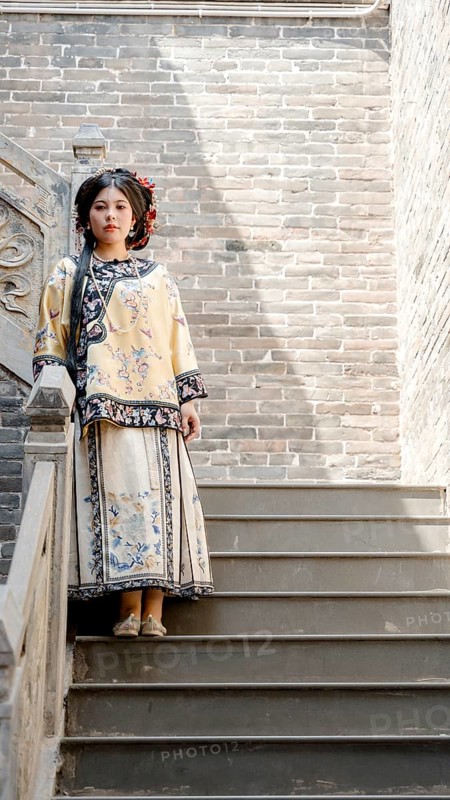 A woman in traditional Qing dynasty clothing stands on stone steps in Pingyao, China, looking at the camera. Her attire features intricate embroidery and vibrant colors