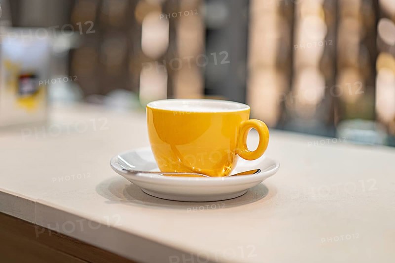 A vibrant yellow coffee cup sits gracefully on a white saucer at an airport cafe, inviting travelers for a refreshing break amidst bustling terminals and waiting passengers