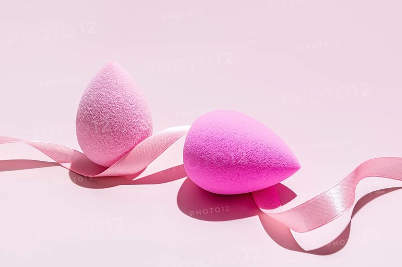 Makeup sponges, a pale and a vibrant pink beauty blender, elegantly intertwined with a silky pink ribbon against a light pink backdrop, illustrating essential beauty tools in a minimalist setting