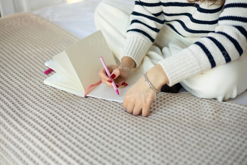 A woman wearing a striped sweater writes in a notebook using a pink pen while sitting on a bed The scene conveys relaxation, creativity, and a cozy atmosphere