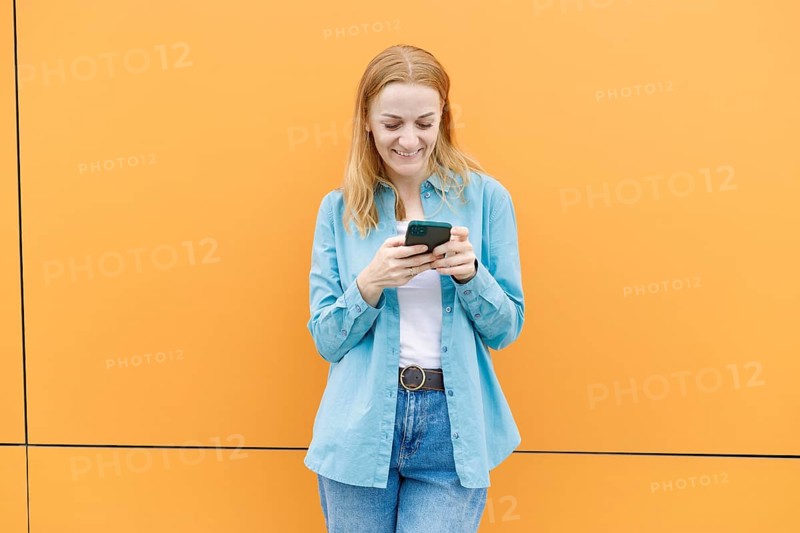A woman in a blue shirt and jeans uses her smartphone while standing against a vivid orange wall in Warsaw The image captures a moment of everyday connectivity and technology