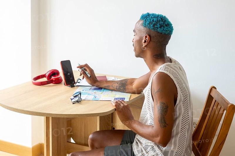 Hispanic LGBTQIA+ man with blue hair planning a holiday trip, sitting at a table with a smartphone, map, and red headphones, embodying pride and creativity