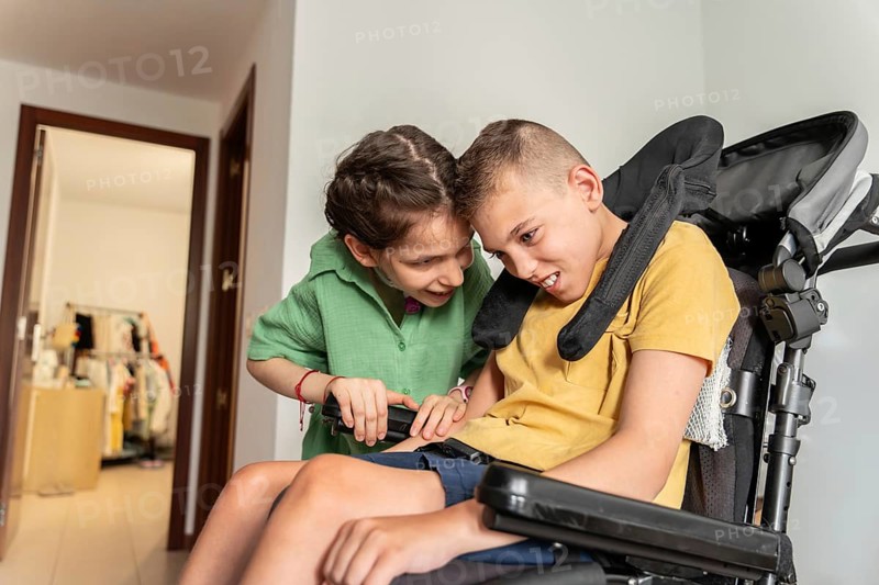 A heartwarming image of two disabled friends, a boy with cerebral palsy and a girl with a tracheostomy, sharing a moment of joy and support at home