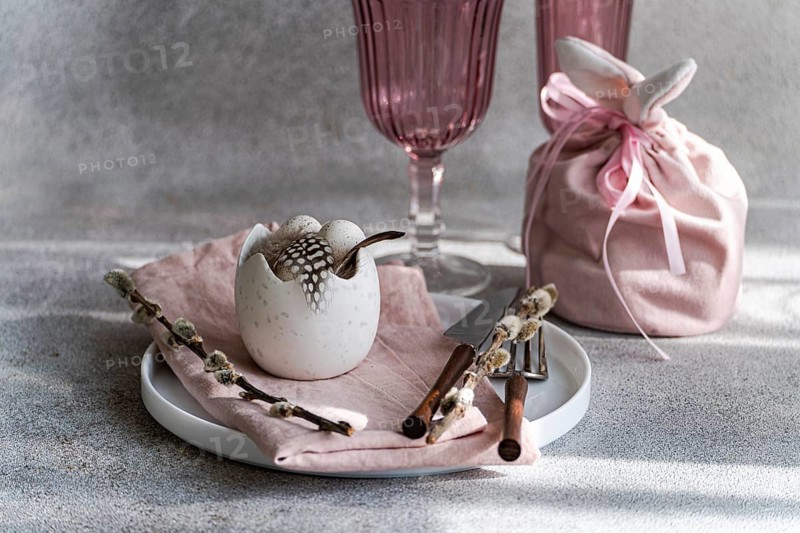 Beautifully arranged Easter table setting featuring a cracked eggshell vase with feathers, surrounded by pink elements and spring decorations. Soft light complements the serene mood