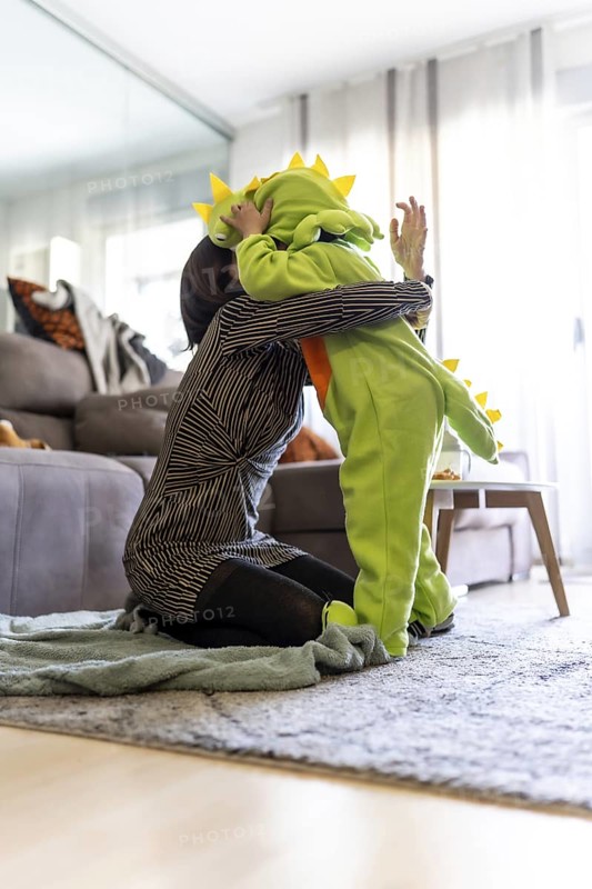 Mother kneeling on carpet and hugging her son wearing a dinosaur costume while playing together in the living room