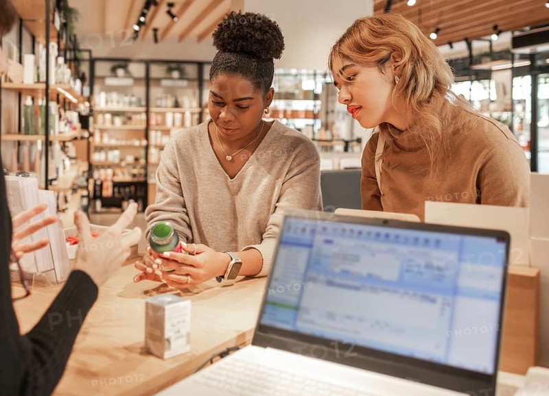 Two women attentively examine a product while receiving expert advice in a modern cosmetics store. A consultant provides guidance, with a laptop and skincare products visible on the counter
