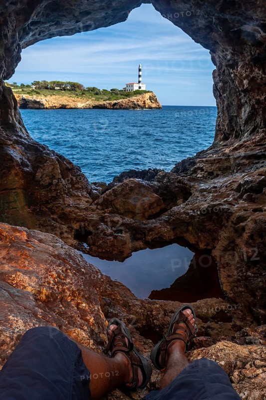 Tourist resting in arc natural cave framing portocolom lighthouse on mallorca island, enjoying scenic coastal view