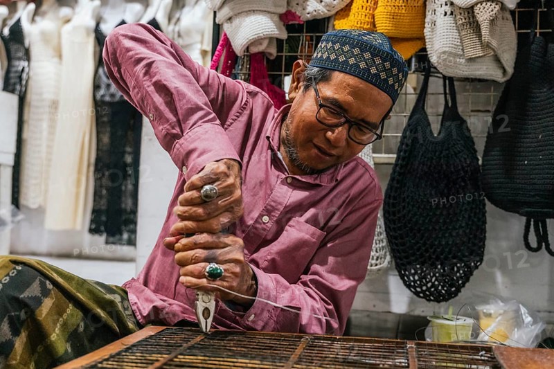 A balinese artisan intensely crafting with pliers and wire, surrounded by vibrant crocheted items in a bustling market. The scene captures creativity, tradition, and craftsmanship