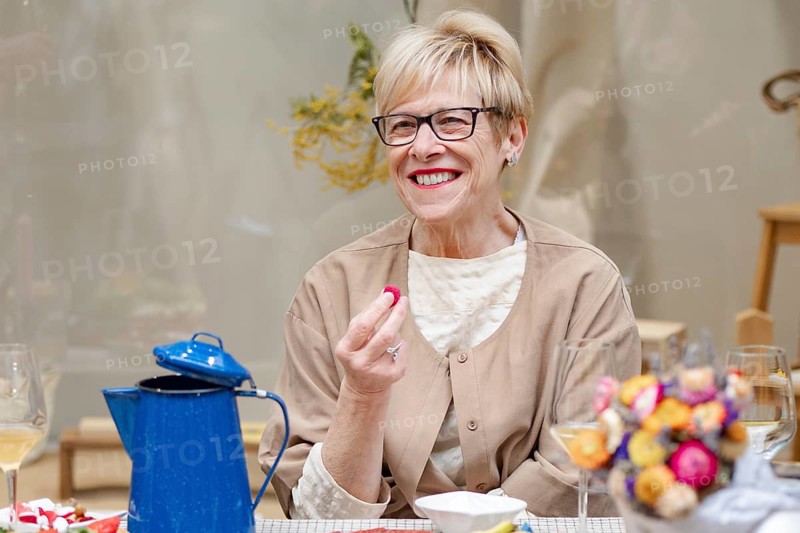 A woman with short hair and glasses is joyfully holding food at a table. A blue teapot and colorful flowers add charm to the cozy dining scene