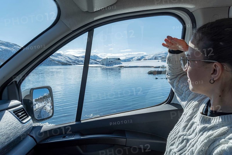 A woman gazes through a car window at the serene, snowy landscape of Botnhamn, Norway, with a calm sea and distant mountains under a bright blue sky
