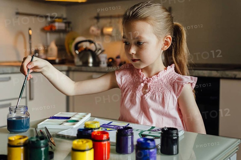 A young girl in a pink dress paints at a kitchen table, surrounded by colorful jars and brushes. The setting is a modern kitchen, emphasizing creativity and imagination