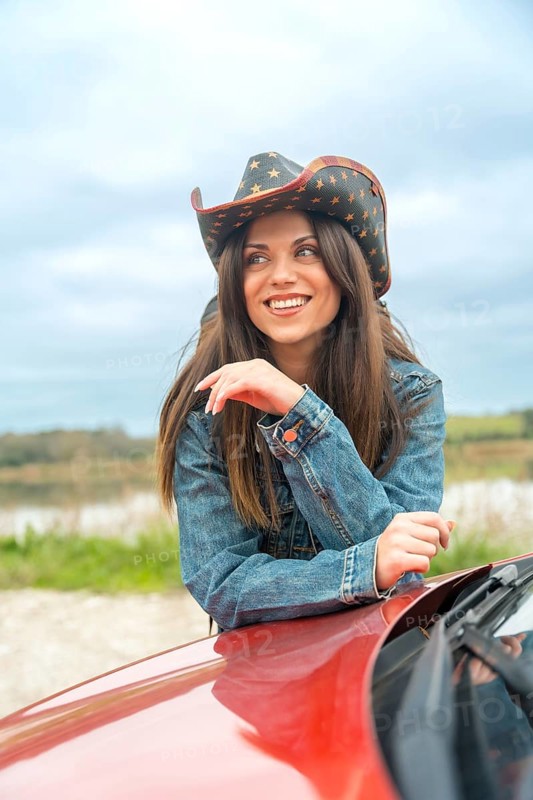 Vertical portrait of a smiling woman with cowboy hat leaning on a car in a road