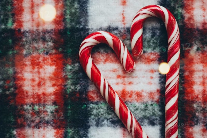 Top view of two red and white striped candy canes arranged in a heart shape against a snowy, textured backdrop, infused with a soft glow of Christmas lights