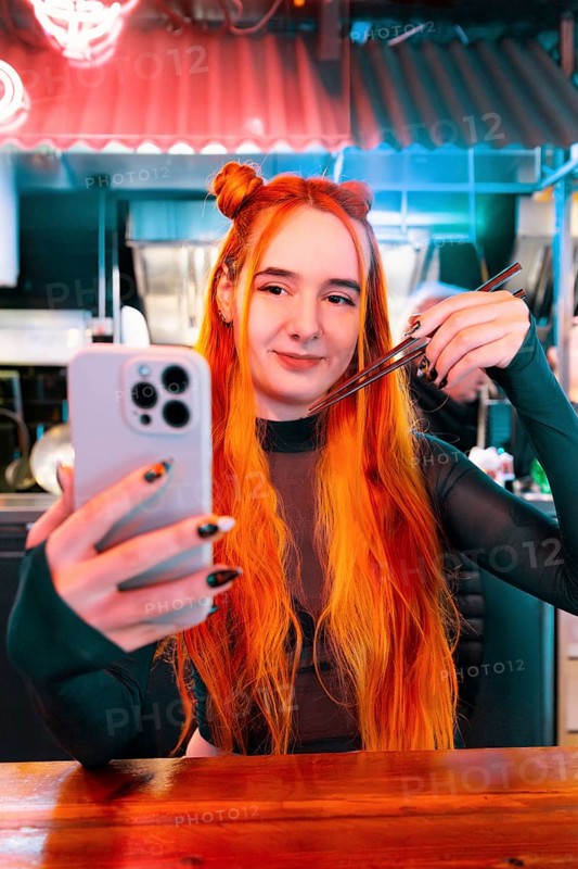 A woman with vibrant hair takes a selfie while visiting an Asian food market She holds chopsticks in hand, capturing the lively atmosphere and diverse culinary experience