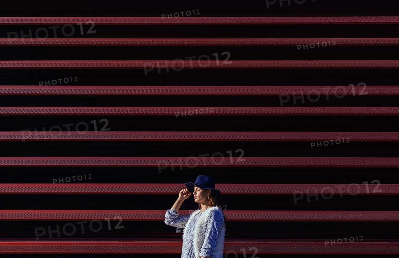 A fashionable woman in a blue hat stands confidently against a striking background of horizontal red and black stripes, creating a bold and modern urban aesthetic