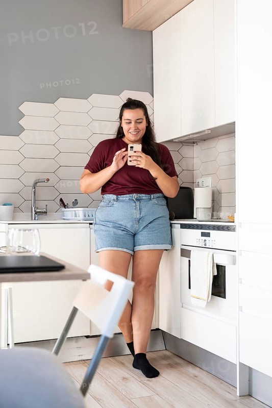 A curvy woman stands in a modern kitchen, smiling while using a smartphone. She leans against the counter, wearing casual clothes. The scene is bright and cozy
