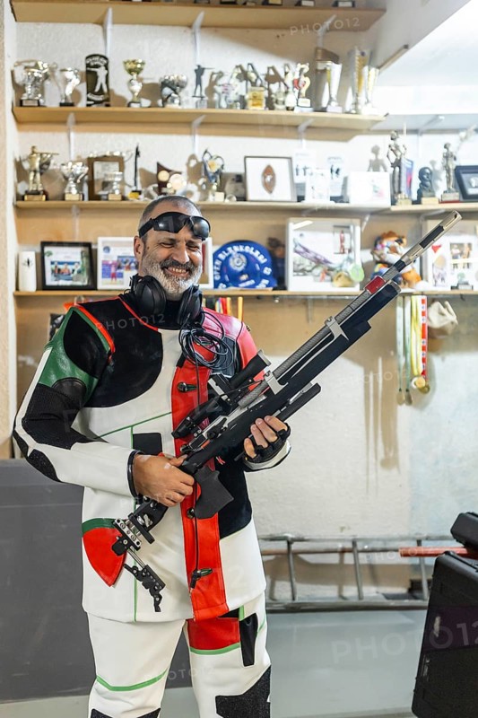 A blind shooter in action, wearing a specialized suit and holding a rifle, standing in front of a shelf filled with trophies, showcasing dedication and achievement