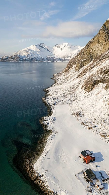 A stunning aerial view of a snow-covered Nordic coastline, showcasing majestic mountains and the tranquil sea under a clear blue sky. A cozy red cabin adds charm to the serene landscape, Kvaløya, Troms, Norway