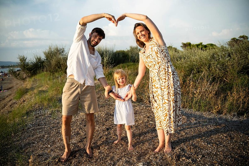 A joyful family forms heart shapes with their arms on a picturesque Italian beach, surrounded by greenery and pebbles, under a sunlit sky