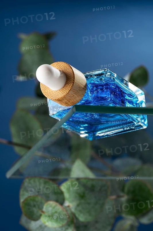A cooling face care serum for men, showcased in a stylish dropper bottle with vibrant blue liquid, contrasted against a blue background surrounded by eucalyptus leaves