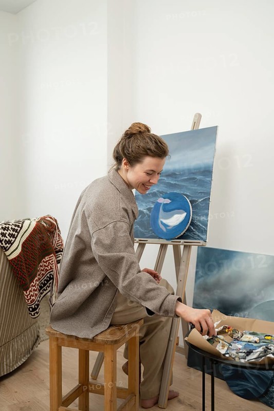 A female artist joyfully paints a whale on a canvas in a cozy home studio. Surrounded by her tools and art supplies, she creates a calming seascape scene