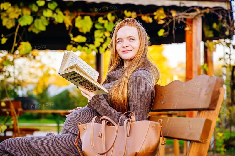 A beautiful pregnant woman with red hair sits on a park bench, enjoying a book. The scene captures the essence of peaceful autumn, highlighted by vibrant foliage