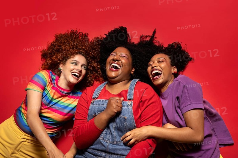 Three friends share a moment of pure joy and laughter, lying on a vibrant red background. Their colorful clothing and genuine expressions highlight the essence of happiness and friendship
