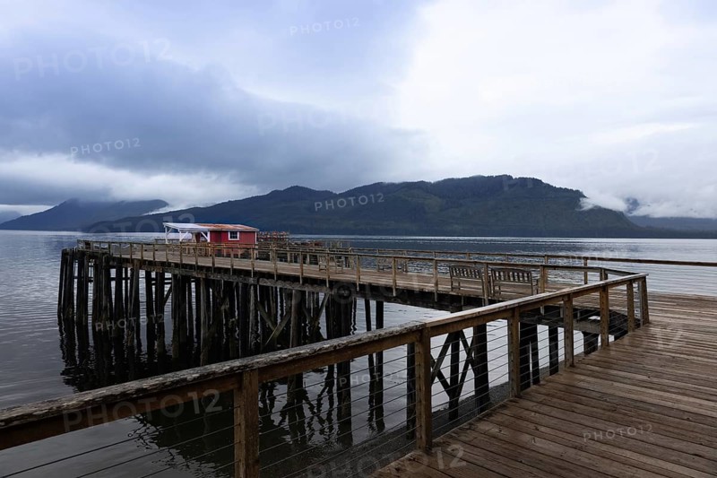 A serene landscape featuring a wooden pier extending into calm waters in Hoonah, Alaska, with a vibrant red building and a dramatic mountain range enveloped by clouds in the background