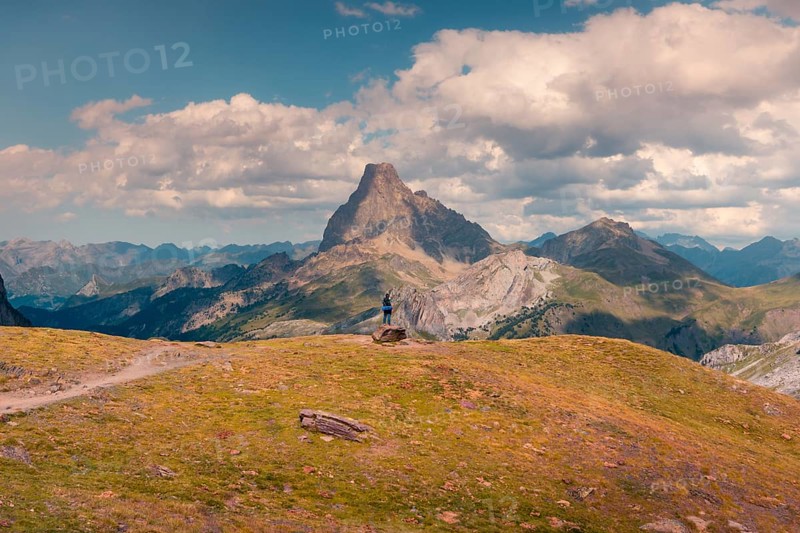 A solitary hiker stands on a rocky hilltop, gazing at towering mountains under expansive skies in the Pyrenees. The lush terrain and dynamic clouds create a stunning natural panorama in summer