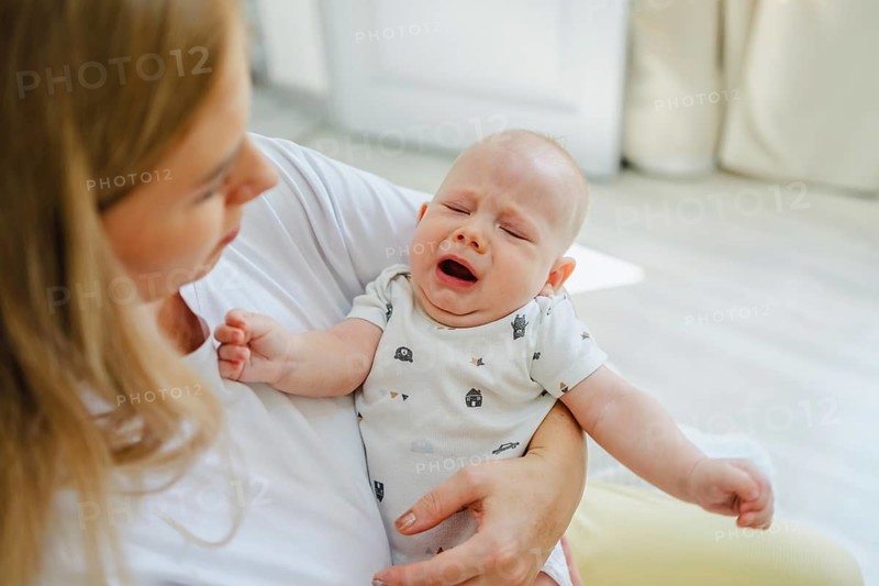A parent gently comforts a crying baby in a warm, bright living room. The scene captures a tender moment of love and care, highlighting the bond between mother and child
