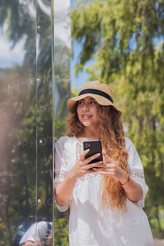 Young American Latin woman enjoying a sunny day outdoors with her smartphone. She wears a stylish hat and white dress, capturing a moment of relaxation and connection