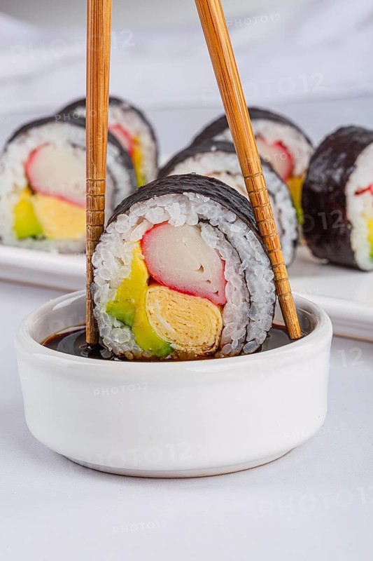 A close up of a sushi roll held by chopsticks, dipping into a soy sauce bowl in white background. Features colorful ingredients like crab stick, avocado, and rolled omelette