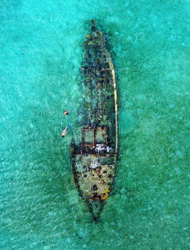 Aerial view of two unrecognizable people, swim around a moss-covered sunken ship in the clear, turquoise waters of the Philippines, highlighting a serene yet adventurous vacation scene
